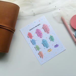 May include: A printed sheet titled "Books of the Bible" with colorful sections listing biblical books. A brown leather journal, a pink pen, and two round, colored paper pads are also visible on a white surface.