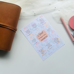 May include: A brown leather journal with a strap closure sits next to a sheet of stickers with floral designs and the text "God is bigger than anxiety." A pink pen and two round sticky notes in pink and blue are also visible.