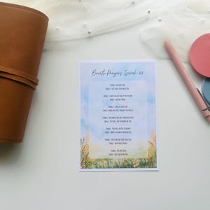 May include: A brown leather journal with a strap closure sits next to a card with the text "Breathe Prayers Isaiah 62". A pink pen and two round coasters, one pink and one blue, are also on the white surface.