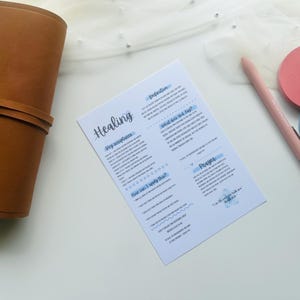 May include: A brown leather journal lies next to a white paper with the word "Healing" in blue script. A pink pen and two round, colored sticky notes are also visible. The journal is closed and has a strap.