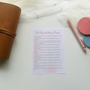 May include: A tan leather journal with a strap, a pink pen, and two circular coasters, one pink and one blue. A printed page with the text "The Proverbs Woman Prayer" is also visible.