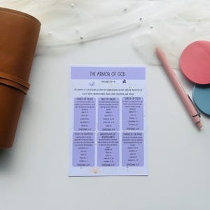 May include: A printed card titled "The Armor of God" with text and scripture references. The card is light purple with white text. A brown leather journal, a pink pen, and two round sticky notes in pink and blue are also visible.