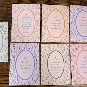 May include: A set of seven rectangular cards featuring floral borders and central oval frames containing religious text. Each card displays a unique botanical pattern in pastel colours, including green, orange, blue, purple, and pink, with a small heart icon.