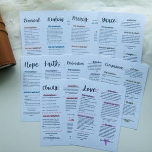 Puede incluir: Tarjetas blancas con títulos escritos a mano como "Renovación", "Curación" y "Misericordia" dispuestas sobre una superficie blanca. Cada tarjeta presenta texto y elementos decorativos en negro, azul y rosa. Un diario de cuero marrón es visible a la izquierda.
