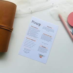 May include: A light-colored paper with the word "mercy" and other text, a brown leather journal, a pink pen, and two round coasters, one pink and one blue. The items are arranged on a white surface.