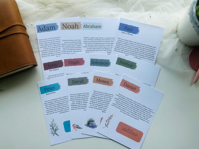 May include: A collection of white cards with names like Adam, Noah, and Abraham, each with a colored banner and text. A brown leather journal is visible on the left. The cards have illustrations of plants and birds.