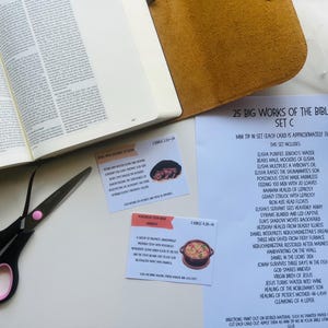 May include: An open Bible with text, a pair of black scissors with pink accents, and a set of Bible study cards. The cards are titled "25 Big Works of the Bible Set C" and include a list of biblical events. A brown leather book cover is also visible.