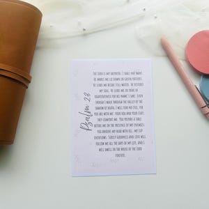 May include: A white card with the text of Psalm 23 in black script, next to a brown leather-bound container, a pink pen, and two circular coasters, one pink and one blue. The text is centered on the card.