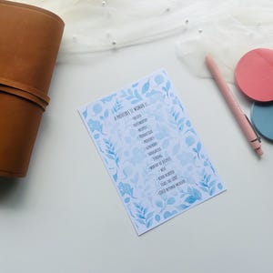May include: A brown leather journal lies next to a card with blue floral accents and text. A pink pen and two round sticky notes, one pink and one blue, are also on the white surface. The card's text reads "A Proverbs 31 Woman Is..."