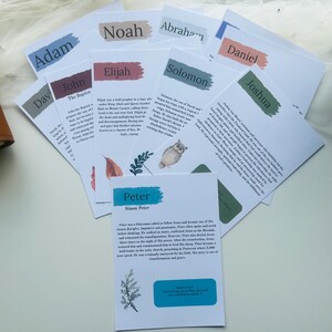May include: A collection of white cards with names like Adam, Noah, and Peter, each featuring a colorful brushstroke and text. Some cards have illustrations of an owl and leaves. A brown leather journal is visible on the left.