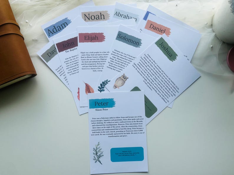 May include: A collection of white cards with names like Adam, Noah, and Peter, each featuring a colorful brushstroke and text. Some cards have illustrations of an owl and leaves. A brown leather journal is visible on the left.