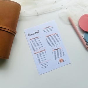 May include: A brown leather journal lies next to a white paper with the word "Renewal" and other text in pink and black. A pink pen and two circular coasters, one pink and one blue, are also on the white surface.