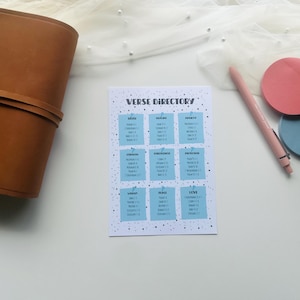 May include: A brown leather journal with a wrap-around closure sits next to a "Verse Directory" card. The card has a light blue background with white text and a pink pen and two circular sticky notes.