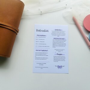 Peut inclure: Une carte blanche avec le mot "Restoration" en écriture cursive, avec du texte et des puces. Un journal en cuir marron, un stylo rose et deux dessous de verre ronds, un rose et un bleu, sont également visibles.