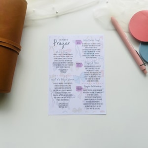 Puede incluir: Una tarjeta blanca con el texto "The Power of Prayer" e información detallada sobre la oración. Un diario de cuero marrón, un bolígrafo rosa y dos posavasos redondos en rosa y azul también están en la imagen.