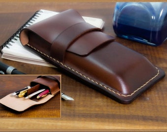 May include: A brown leather pen case with a flap closure and a stitched design. The case is open to reveal multiple pens inside.