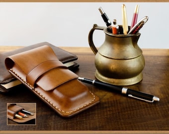 May include: A brown leather pen case with a flap closure and stitching detail. The case is open to reveal a compartment for pens. The case is on a wooden surface with a brass pitcher filled with pens in the background.
