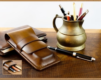 May include: A brown leather pen case with a flap closure. The case is sitting on top of a brown leather notebook. A black pen is lying on the table next to the case. A brass pitcher with a handle is filled with pens.