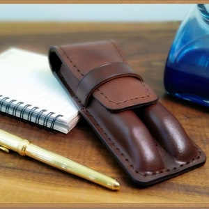 May include: A brown leather pen case with two slots for pens. The case has a flap that closes with a snap. A gold pen is lying on a wooden surface next to the case.