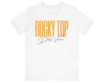 May include: A white short-sleeved t-shirt featuring the text ROCKY TOP in bold yellow capital letters with a subtle pink drop shadow, followed by the text Dolly's Version in a black cursive script font centered on the front of the garment.