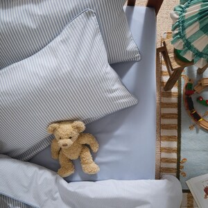 May include: A light blue bed set with striped pillowcases and a matching flat sheet. A small, tan teddy bear rests on the bed. A small wooden train set and a green and white checked pillow are also visible.