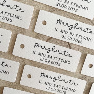 Può includere: Primo piano di diversi tag rettangolari bianchi con un piccolo foro nella parte superiore. Ogni tag ha il nome "Margherita", il testo "IL MIO BATTESIMO 21.09.2025" stampato in nero e una superficie strutturata.