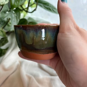 May include: A small, handmade ceramic bowl with a glossy, iridescent glaze in shades of green, blue, and brown. The bowl has a reddish-brown base and is held in a hand. The bowl is likely used for serving small portions of food or as a decorative item.