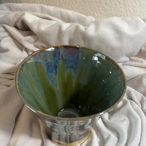 Op de afbeelding: Een handgemaakte keramische kom met een uitlopende rand. De binnenkant heeft een mix van groene en blauwe tinten, terwijl de buitenkant een getextureerde, grijze afwerking heeft. De rand is geaccentueerd met een bruine band. De kom staat op een lichtbeige basis.