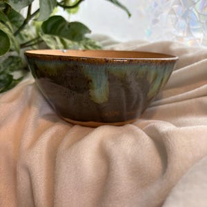 May include: A handmade ceramic bowl with a dark green and teal glaze. The bowl has a wide, shallow shape with a brown rim and base. The bowl is sitting on a soft, white surface.