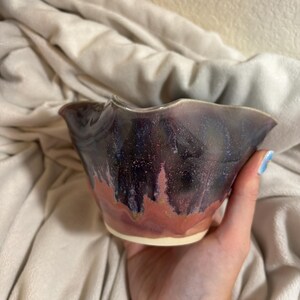 May include: A handmade ceramic bowl with a unique glaze. The bowl features a dark purple and red color scheme with a textured surface. The rim has a wavy design. The bowl is held in a hand.