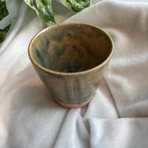 Puede incluir: Una taza de cerámica hecha a mano con un esmalte único. La taza presenta una mezcla de tonos terrosos, incluyendo verdes, marrones y toques de azul. La base tiene un tono rosa pálido. La taza está sobre un fondo blanco texturizado.