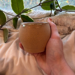 Peut inclure: Une petite tasse en céramique beige mouchetée, de forme arrondie, avec un émail vert clair lisse qui coule sur le bord supérieur. La tasse présente une finition mate et texturée sur le corps extérieur, avec une section centrale légèrement plus large.