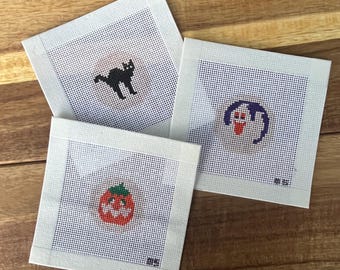 May include: Three square needlepoint canvases featuring Halloween designs. One canvas shows a black cat, another a white ghost with a purple outline, and the third a small orange pumpkin with a green stem. Each design is centered on white mesh.