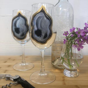 May include: Two clear wine glasses with a gold rim and a gray and black abstract design. A clear bottle and a small vase with purple flowers are behind the glasses. A corkscrew is in the foreground.