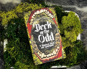 May include: A book titled "Deck of the Odd: A Handcut Tarot and Guidebook" with a colorful illustration of a mouth with teeth on the cover. The book is lying on a bed of green moss.