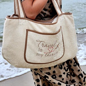 Könnte beinhalten: Eine beige Tote Bag mit braunem Besatz und einer Tasche auf der Vorderseite. Die Tasche ist mit dem Text "Travel is my therapy" in braunem Garn mit einem roten Herzen und einem Flugzeugsymbol bestickt.