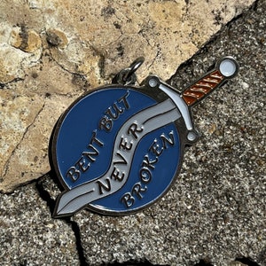 May include: A blue enamel pin with a silver sword design. The sword is bent but the text on the pin reads "Never Broken".