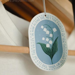 May include: A blue and white oval air freshener with a lily of the valley illustration and the text "The lily of the valley, in him alone see... To my soul. The fairest of ten thousand."  The air freshener is hanging from a wooden hanger.