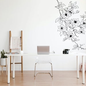 May include: A white desk with a laptop, a camera, and a plant on it. A white ladder with a pink and white blanket draped over it is in the background. A black and white floral decal is on the wall behind the desk.