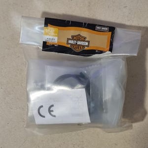 Op de afbeelding: Een doorzichtige plastic zak met motorfietsonderdelen. De zak heeft een label met het Harley-Davidson-logo en de tekst "MOTOR HARLEY-DAVIDSON CYCLES". Het label bevat ook de tekst "H.D. 011 5343CMC011".