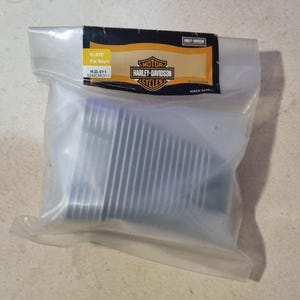 Op de afbeelding: Een doorzichtige plastic zak met een motorfietsonderdeel. De zak heeft een zwart en geel label met het Harley-Davidson logo en de tekst "MOTOR HARLEY-DAVIDSON CYCLES". Het label bevat ook de tekst "FL5TF Fat Boy®".