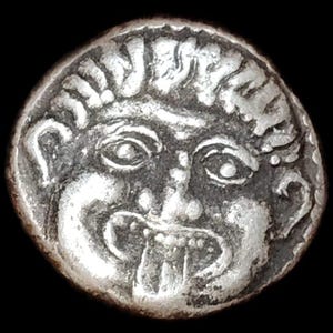 May include: A silver coin featuring a stylized face with large eyes, a wide mouth, and detailed hair. The coin has a worn appearance, suggesting age, and is set against a black background. The design is centered and well-defined.