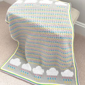 May include: A gray crocheted blanket featuring white clouds with pink, blue, and yellow raindrops. The blanket has a colorful border and is draped over a neutral-colored surface. The blanket is a handmade item.
