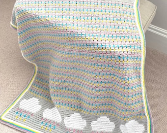 May include: A grey crocheted blanket with white clouds and pink, blue, and yellow raindrops. The blanket has a colourful border and is draped over a neutral surface. The blanket is a handmade item.
