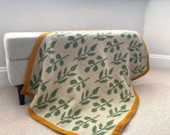 May include: A knitted blanket with a leaf pattern in green and beige, edged with a mustard yellow border. The blanket is draped over a bench. The design features repeating green leaves on a beige background.