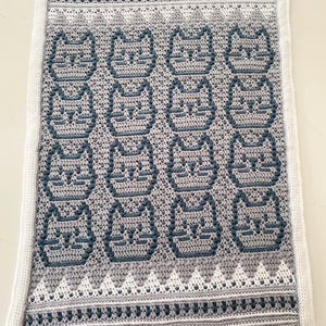 May include: A crocheted blanket with a repeating pattern of cat faces in grey and blue. The blanket has a white border and a decorative border with a geometric design. The design has a handmade aesthetic, perfect for a cosy home.