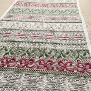 May include: A rectangular, crocheted blanket with a repeating pattern of bows, leaves, and trees in shades of gray, green, and red. The blanket has a white border and is likely handmade. The design is reminiscent of a holiday theme.