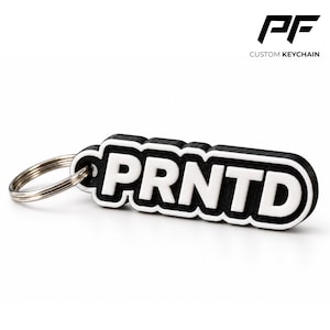 May include: A black and white keyring featuring the text PRNTD in bold, raised white letters on a black background. The keyring has a rounded rectangular shape with a thick white border and includes a silver metal split ring for attachment.