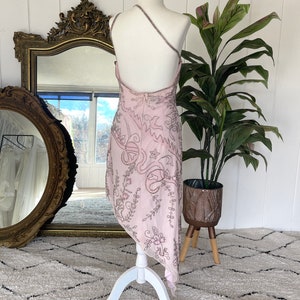 May include: A pink, one-shoulder dress with a floral pattern and silver embroidery. The dress is on a white dress form and has a flowing, asymmetrical hemline.