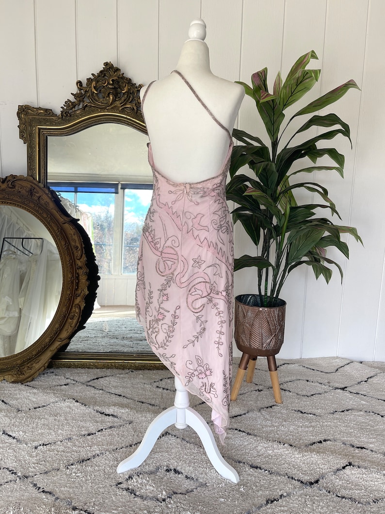 May include: A pink, one-shoulder dress with a floral pattern and silver embroidery. The dress is on a white dress form and has a flowing, asymmetrical hemline.